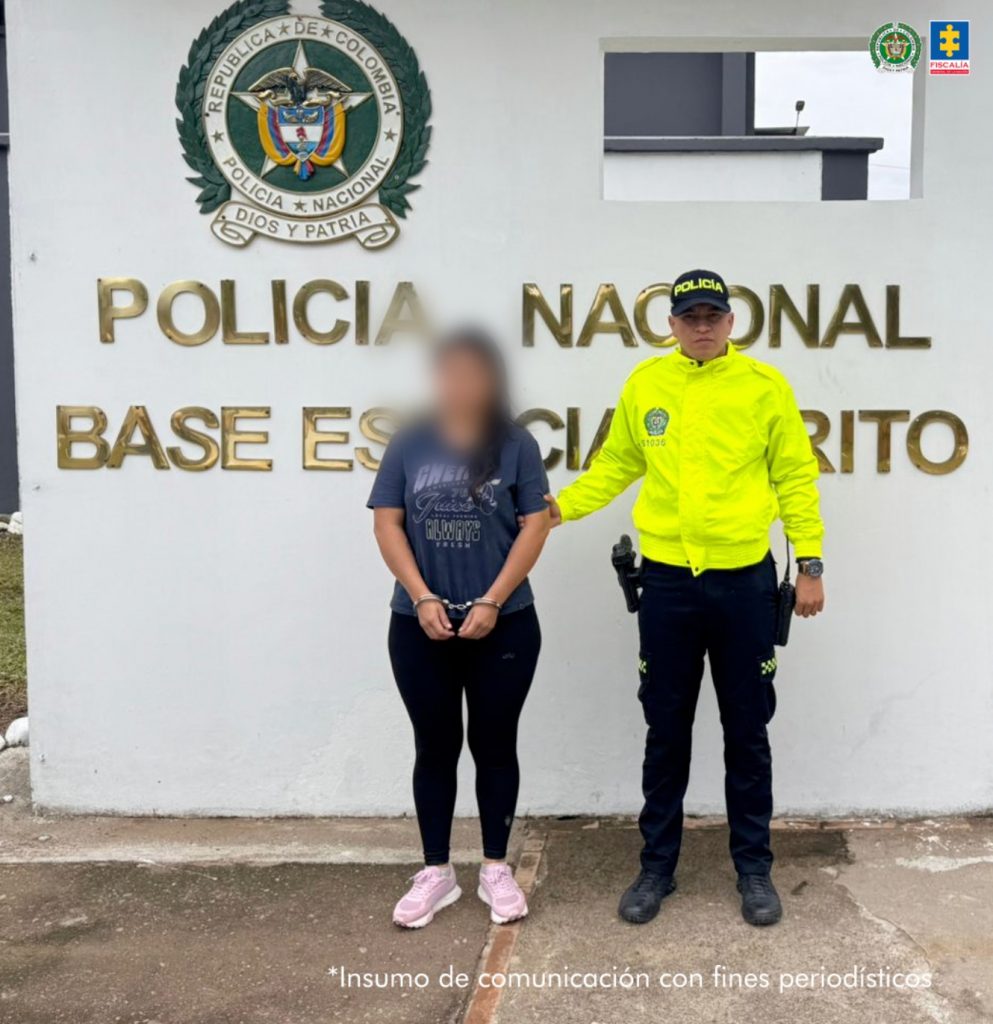 En la foto se observa a la capturada custodiada por un uniformados de la Policía Nacional. Detrás hay un muro con blanco con distintivos de la Policía Nacional.