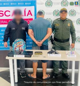 Se visualiza al capturado junto a investigador del CTI y un uniformado de la Policía Nacional. Frente a ellos una mesa con los elementos y sustancia incautada. Detrás de ellos los banners que identifican a la Fiscalía General de la Nación y a la Policía Nacional.