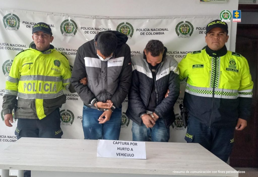 en la imagen se puede observar a los dos capturados en medios de dos agentes de policia, en frente de ellos se ve una mesa con un letrero que dice captura por hurto de vehiculo. detras de ellos se ve un baking de la policia
