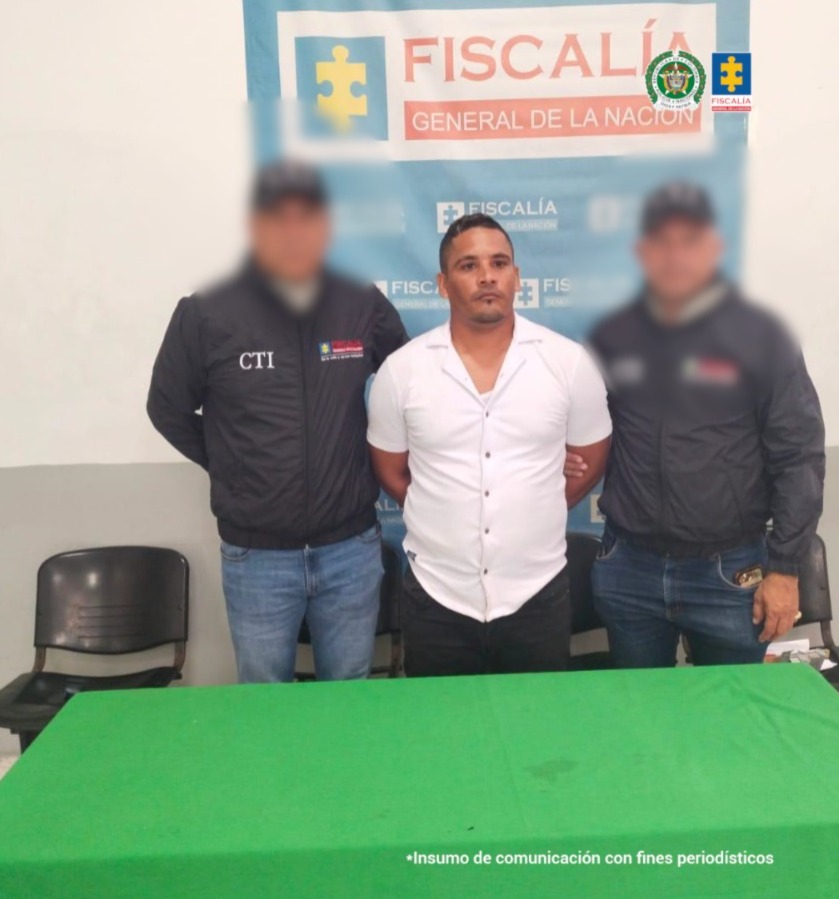 En la imagen se ven dos uniformados en compañía del capturado quien tiene una camisa blanca. Los rostros de los uniformados están difuminados. 