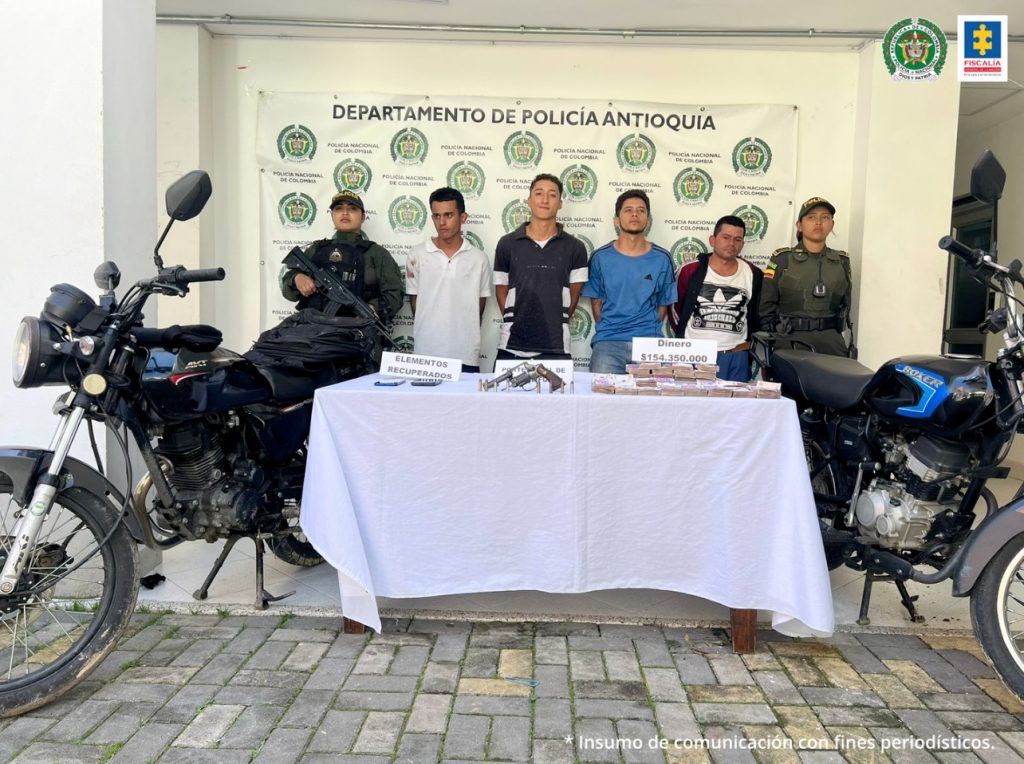 En la imagen están dos Policías de sexo femenino y en medio los 4 procesados con una mesa en la que se encuentran un arma de fuego, dinero en efectivo, dos celulares y dos motocicletas; elementos incautados durante la captura.