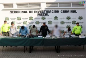 En la fotografía se aprecia en hilera, con la cabeza agachada y esposados entre sí, a los presuntos integrantes de una banda, dedicada al hurto violento. Entre ellos hay una mujer. A los extremos hay uniformados de la Policía Nacional. Delante de ellos hay una mesa larga con mantel verde y sobre ella se alcanzan a visualizar varios elementos como armas, munición, relojes, dinero en efectivo, celulares, un portátil, entre otros. Detrás de ellos hay un pendón institucional de la Policía.