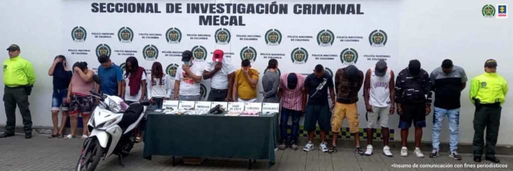- En la fotografía aparecen 13 personas, entre ellas 4 mujeres, capturadas junto a dos agentes de la Policía Nacional. En la parte posterior se ve una mesa con varios elementos incautados y una motocicleta.