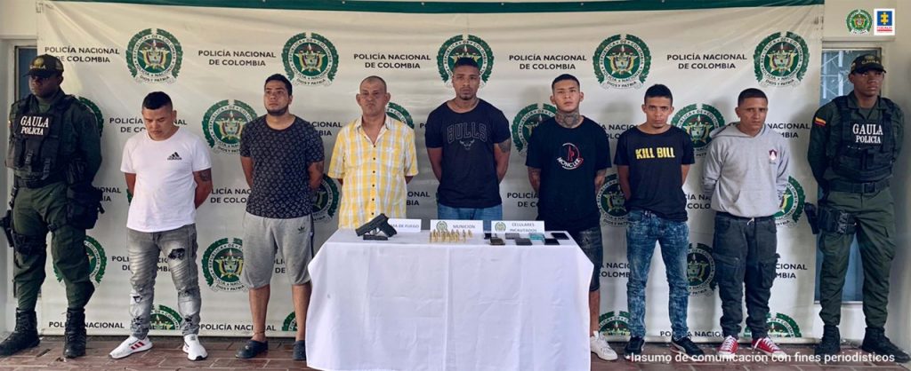 En la fotografía aparecen 7 presuntos miembros de la banda Los Vigilantes junto a dos unidades del Gaula de la Policía Nacional. En la parte posterior se ven una mesa con un arma, municiones, 7 teléfonos celulares y un banner de la Policía Nacional.