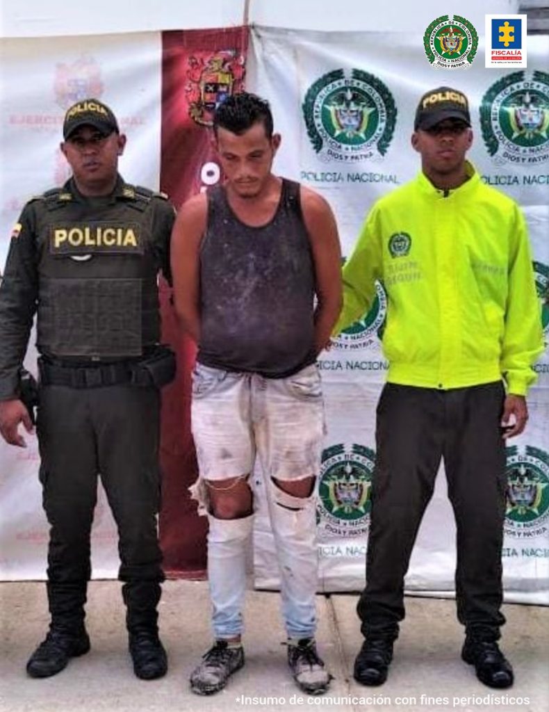 El capturado está parado con sus manos esposadas a su espalda y está custoridado por dos hombres uniformados de la Policía nacional.