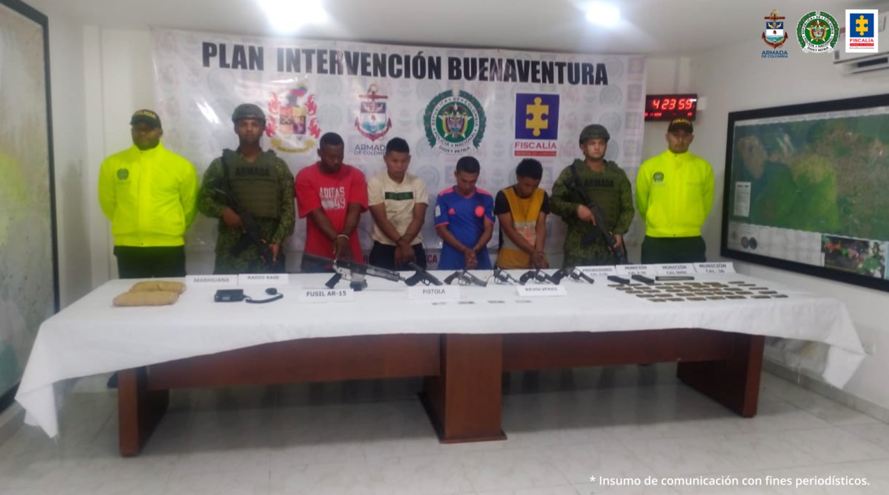 En la fotografía se observan los cuatro judicializados con dos agentes de la Sijin y dos unidades de la Armada Nacional. En la parte posterior se observa una mesa con armas, municiones, proveedores, cédulas, un radio de comunicaciones, y marihuana
