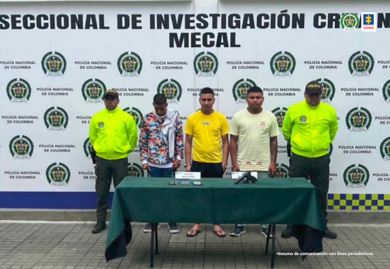 En la fotografía se observan los tres capturados junto con dos agentes de la Policía Metropolitana de Cali. En la parte posterior se observa una mesa con un arma de fuego y dos teléfonos celulares.