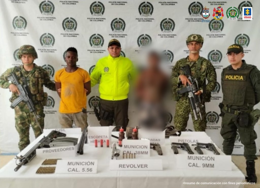 En la fotografía aparecen los dos capturados junto a dos agentes de la Policía Nacional, un infante de marina y un soldado del Ejército Nacional. En la parte posterior se observa una mesa con armas de fuego, municiones, explosivos artesanales y una mochila.