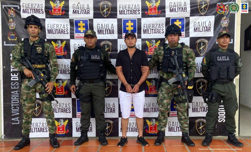 Está el capturado en bermudas, esposado manos al frente, custodiado por uniformados de Policía y Ejército. Detrás banner de grupos Gaula Policía, Ejército y Fiscalía.