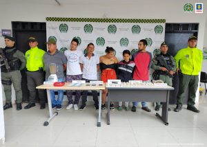 La foto muestra a los cinco acusados ​​de pie, incluida una mujer.  A ambos lados de ellos se encuentran uniformados de la Policía Nacional.  Frente a ellos se encuentra una mesa con un mantel verde y sobre ella varios elementos incautados como marihuana, licuadora, dinero en efectivo y estupefacientes.  Detrás de ellos hay una pancarta institucional policial.