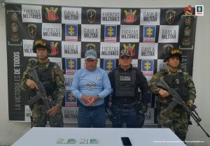 Capturado de frente, esposado y custodiado por Investigadores del CTI y militares del Ejército Nacional. Detrás hay un banner de grupo Gaula Militar.