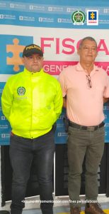 El capturado de frente, custodiado por un Policía y detrás banner de la Fiscalía.