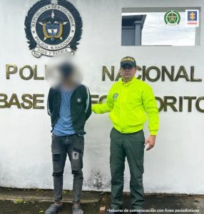 Está de frente el capturado esposado y custodiado por dos uniformados de la Policía, detrás banner de la Policía Nacional.