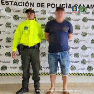 capturado esposado manos atrás custodiado por un uniformado de la Policía Nacional, detrás el banner de la Policía Nacional.