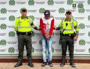 capturado esposado con gorra manos adelante, custodiado por dos policías. Detrás banner de la Policía.