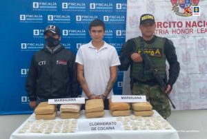  está el capturado de frente esposado custodiado por un investigador de CTI de la Fiscalía y un militar, delante de ellos una mesa con material incautado y detrás banner de la Fiscalía y de Ejército.