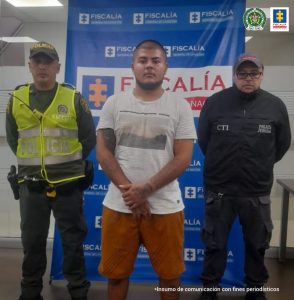 capturado de frente custodiado por investigador del CTI de la Fiscalía y policía. Detrás banner de fiscalía.