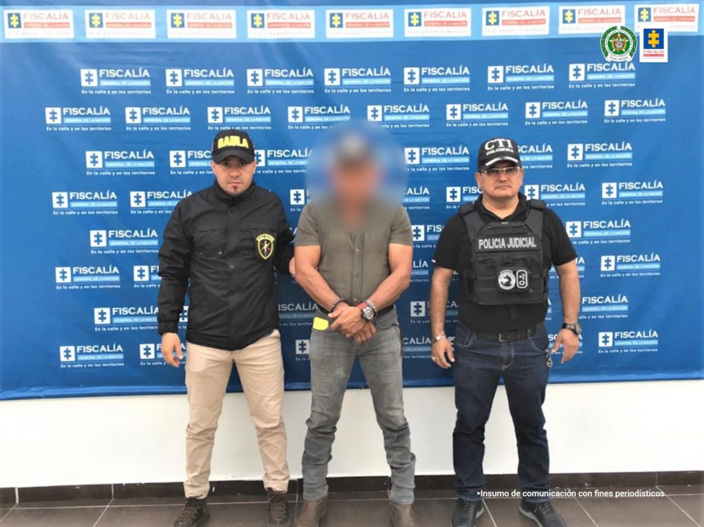 🔼 El capturado está de frente a la cámara con sus manos esposadas, como custodios están un servidor del CTI de la Fiscalía y uno del Gaula Militar, detrás de ellos, un pendón de la Fiscalía general de la Nación.