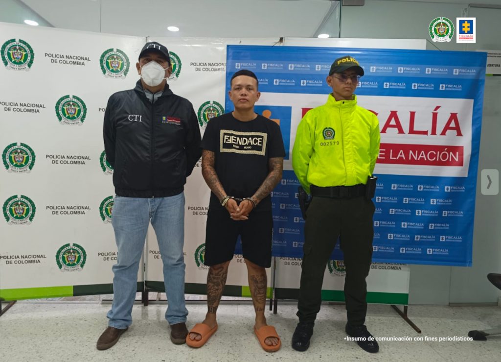 capturado custodiado por investigador CTI de la Fiscalía y un policía. Detrás banner de Policía Nacional y Fiscalía.