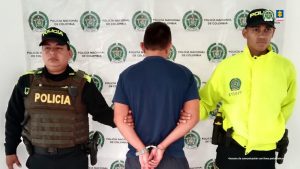 El capturado está de espaldas y rodeado por policías. Detrás banner de la Policía Nacional.