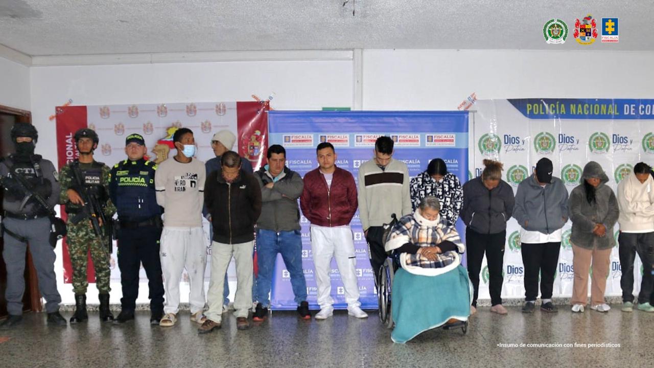 Los capturados aparecen de pie, en línea recta, algunos con las manos adelante y otros con las manos atrás. Tienen a miembros de la Policía, el Ejército y el CTI al lado y banner de las tres instituciones atrás. Una de los capturados está en silla de ruedas. 