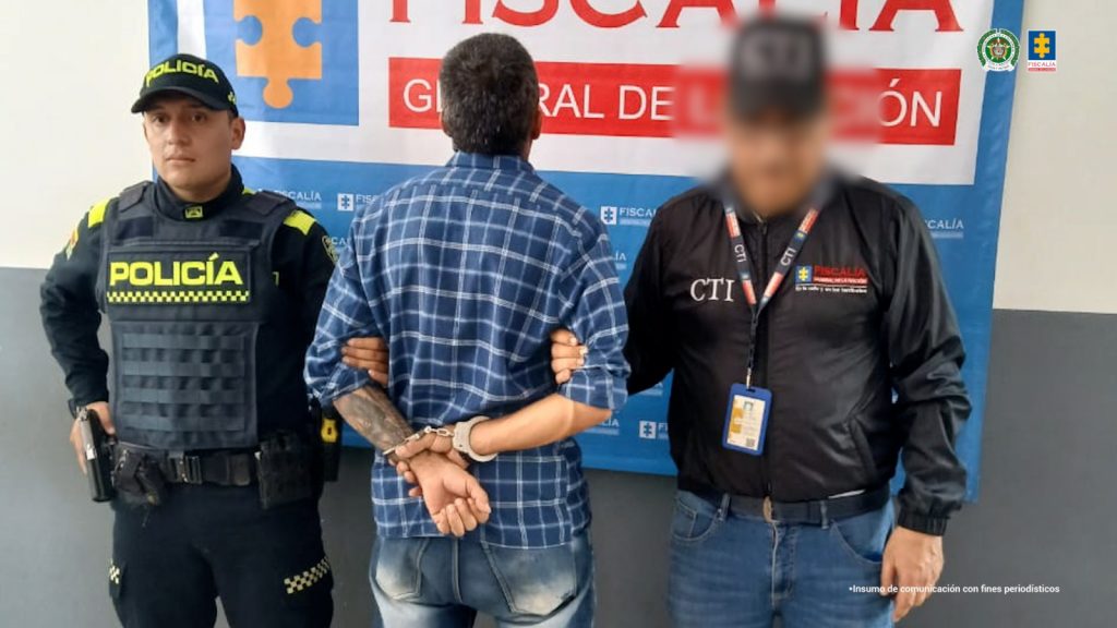 capturado de espaldas custodiado por investigador del CTI de la Fiscalía y un policía. Detrás banner de la Fiscalía.