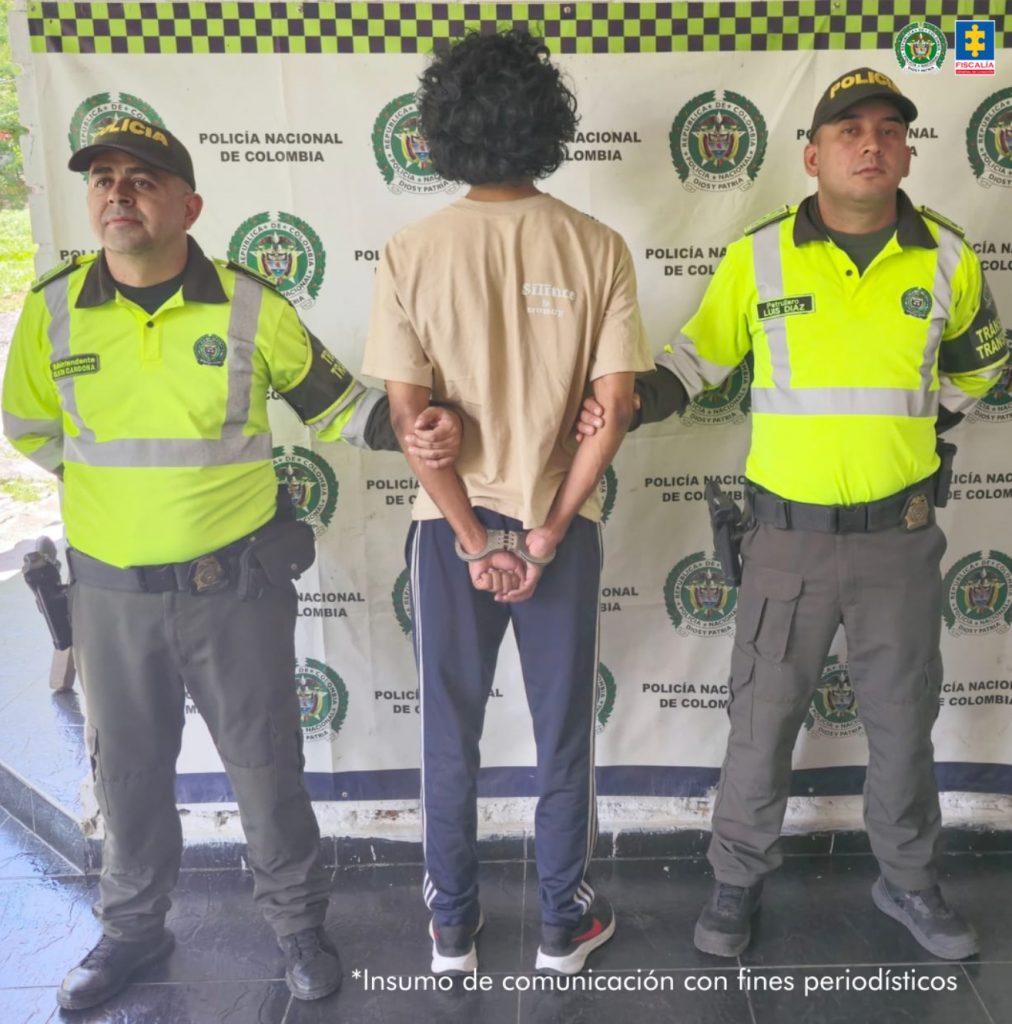 En la imagen se ve a un hombre esposado, de espaldas, custodiado por dos policías uniformados de la Policía Nacional de Colombia. El detenido está en el centro de la foto, vestido con una camiseta beige clara y pantalones deportivos oscuros con franjas blancas a los lados. Ambos policías llevan uniformes verde fluorescente con detalles reflectivos y pantalones oscuros, y están posicionados a cada lado del hombre, sujetándolo por los brazos.De fondo, hay un pendón institucional con los logos de la Policía Nacional de Colombia y de la Fiscalía General de la Nación. En la esquina inferior derecha de la imagen, hay un texto que dice: "Insumo de comunicación con fines periodísticos".