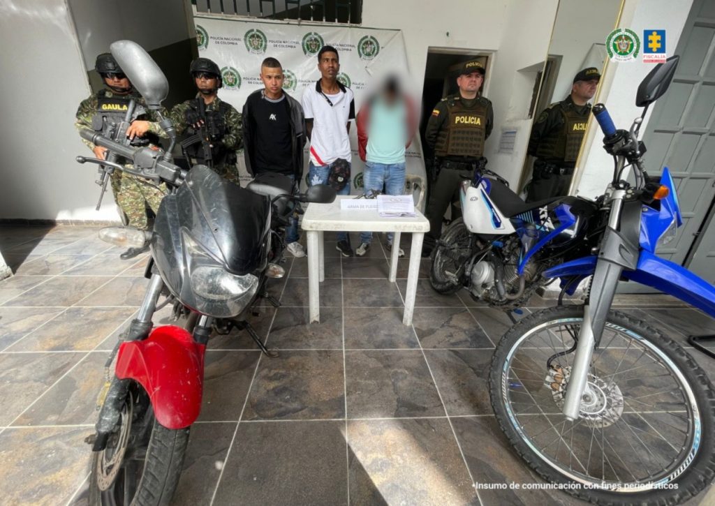 Capturados de frente custodiados por policías, delante de ellos material incautado. Detrás banner de la Policía Nacional.