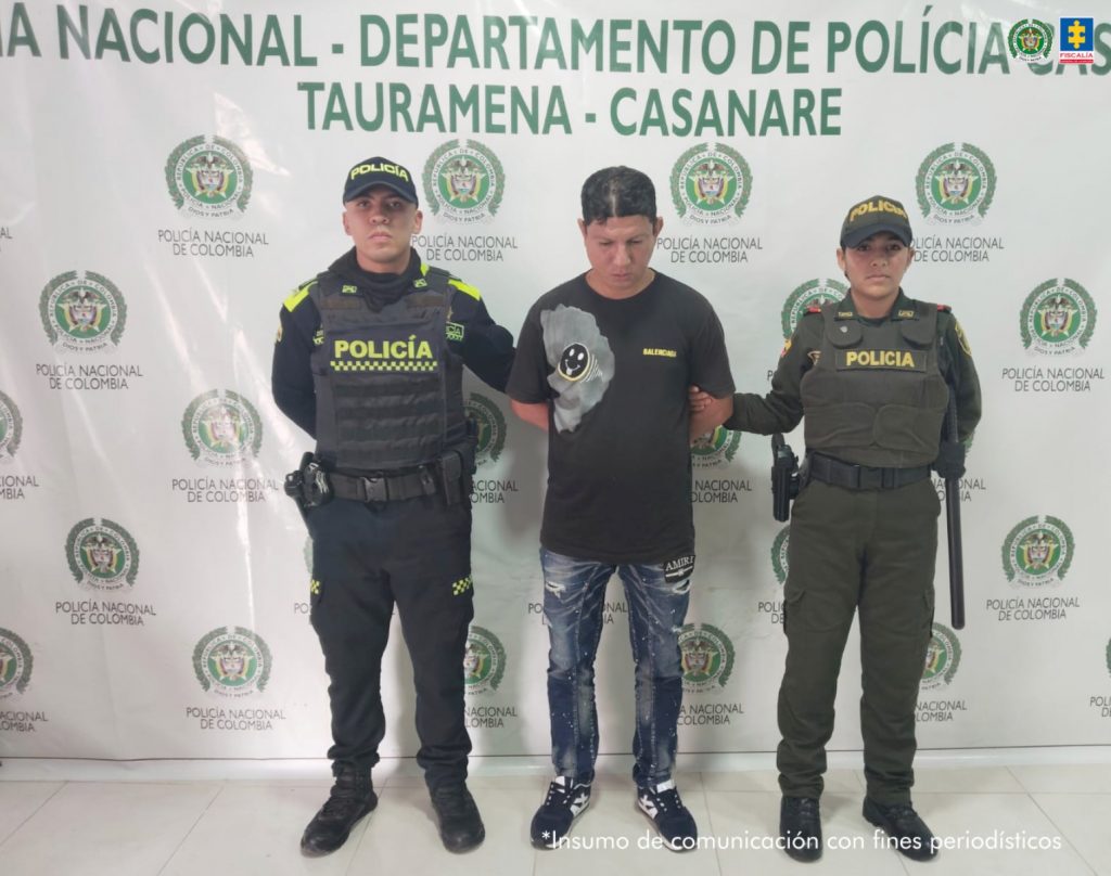 capturado de frente custodiado por dos policías. Detrás banner de la Policía Nacional.