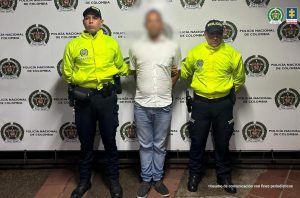 capturado de frente custodiado por policías. Detrás banner de la Policía Nacional.