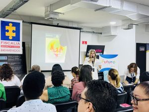 En la imagen se observa una sala de conferencias o aula donde se está llevando a cabo una presentación o taller.Hay varias personas sentadas de espaldas, prestando atención a una mujer que está de pie frente a ellos con un micrófono y una hoja en la mano, quien parece ser la conferencista o facilitadora del evento. A su lado, otra mujer está escribiendo o registrando algo sobre una mesa.  En la pared del fondo se proyecta una diapositiva con un diseño que incluye una figura de un ave fénix y el texto “Renaciendo”, junto con la palabra “Santander”.  A la izquierda se ve un cartel con el logotipo y nombre de la Fiscalía General de la Nación y la palabra “Bienvenidos”. También hay otros carteles con mensajes institucionales, entre ellos uno que dice “Futuro Colombia”.