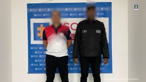 En la fotografía se aprecia de pie y con los brazos atrás al instructor deportivo. Viste camiseta blanca con estampado de colores rojo y negro en la parte superior, incluyendo del cuello; así como pantalón negro. Al lado izquierdo de él hay un servidor del CTI y detrás de ellos un pendón institucional de la Fiscalía.