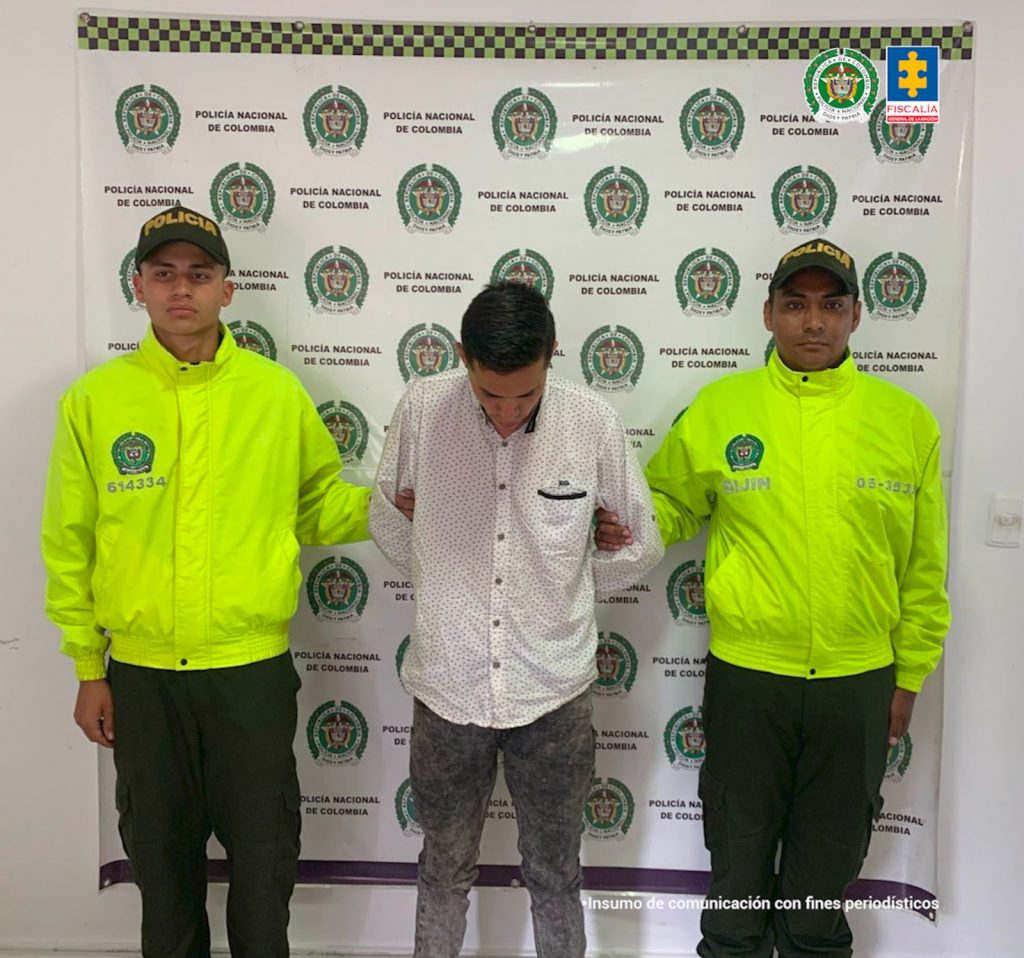 en la fotografia se puede observar al detenido entre dos agentes de policia, detras de ellos se encuentra un baking de la policia nacional