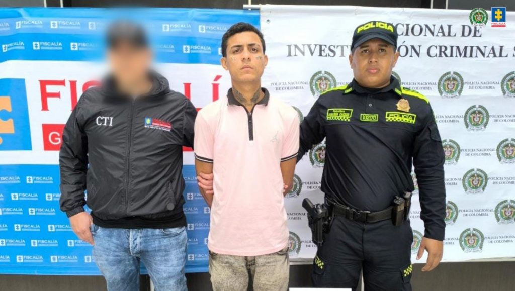 e visualiza al capturado junto a un investigador del CTI y un uniformado de la Policía Nacional. Detrás de ellos los banners que identifican a la Fiscalía General de la Nación y a la Policía Nacional.