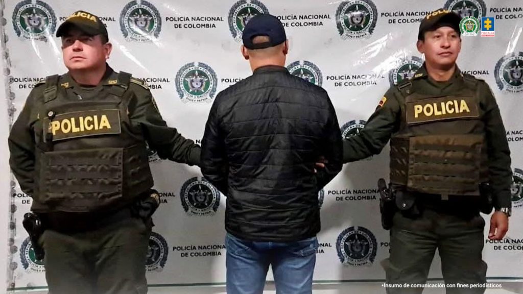 Se visualiza al capturado de espalda junto a dos uniformados de la Policía Nacional. Detrás se ubica el banner que identifica a la Policía Nacional.