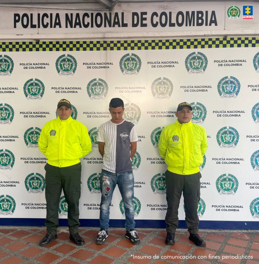 Se observa al capturado de frente. A cada costado un funcionario de Policía Nacional y atrás un pendón de Policía Nacional