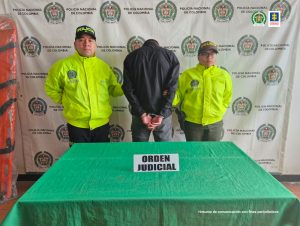 Se observa al capturado de espaldas y a cada costado un funcionario de Policía Nacional. Al frente una mesa con el aviso ORDEN JUDICIAL. Atrás un pendón de Policía Nacional