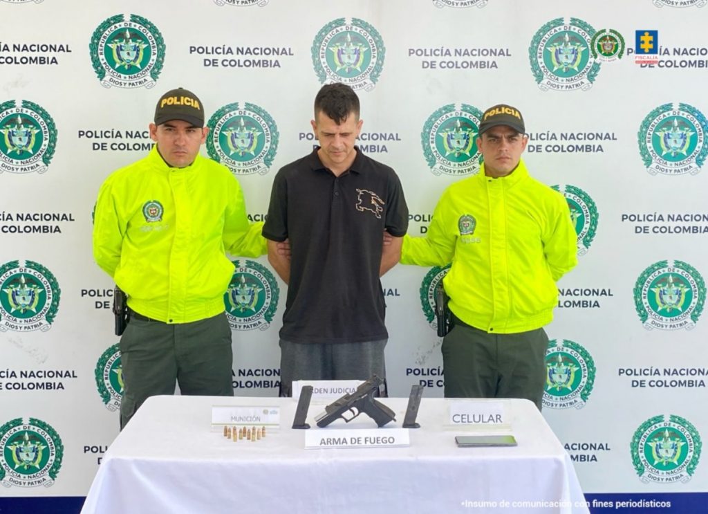 Se observa al capturado de frente.  A cada costado un funcionario de Policía Nacional, atrás un pendón de Policía Nacional y adelante una mesa con el material incautadoPIE DE FOTO: Jhon Andrés Silva Patiño fue detenido  en el barrio Veracruz de la comuna Villasantana de Pereira (Risaralda).
