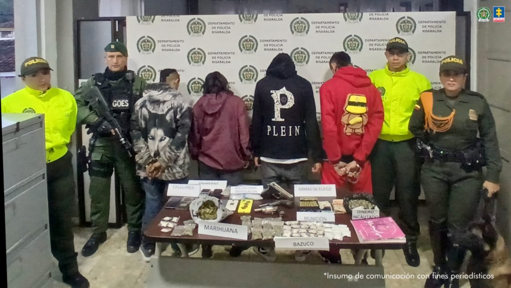 Se observan cuatro capturados en fila y de espaldas. A cada costado los custodian dos funcionarios de la Policía Nacional. A uno de los uniformados lo acompaña un canino de Policía Nacional. Adelante una mesa en la que se exponen los elementos incautados durante las diligencia de allanamiento. Atrás, un pendón de Policía Nacional.