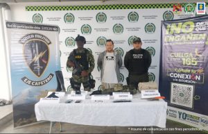 Se observa el capturado de frente.  A un costado un uniformado del Ejército Nacional y al otro uno de Policía Nacional.  Adelante una mesa con el material incautado.  Atrás pendón de Policía Nacional y del Ejército Nacional