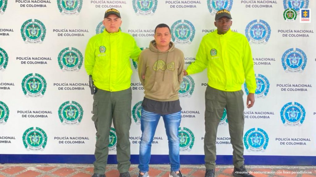 Se observa al capturado en el centro y a cada costado un uniformado de Policía Nacional. Al fondo un pendón de Policía Nacional