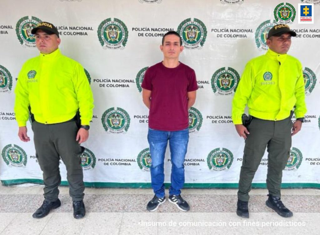 en la imagen se ve una persona detenida en custodia de dos uniformados de la Policía Nacional. Detrás suyo un backing institucional.