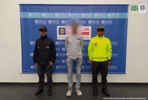Se observa al capturado en el centro, a un costado un funcionario del CTI y al otro uno de Policía Nacional. Atrás un pendón de la Fiscalía General de la Nación.