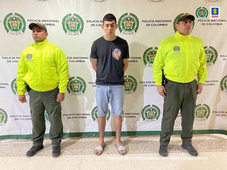 En la imagen se ve una persona detenida bajo custodia de dos uniformados de la Policía Nacional. Detrás suyo un backing institucional.