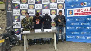 Se observan dos hombres capturados, a un costado un funcionario del CTI y al otro uno del ejército nacional. Atrás un pendón de la Fiscalía general de la Nación y otro del Ejército Nacional. Al frente una mesa con material incautado y a un costado una moto.