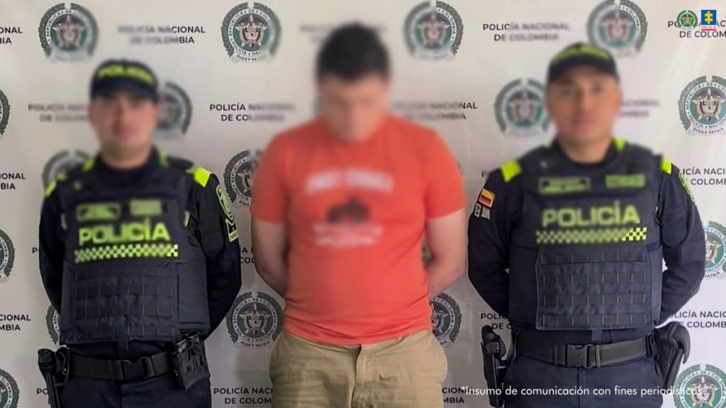 Se observa al capturado de frente y a cada costado un uniformado de la Policía Nacional. Atrás, un pendón de Policía nacional