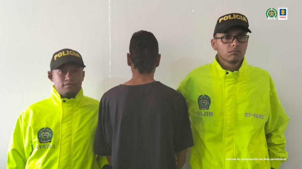 Se observa al detenido de espaldas y en cada costado un funcionario de la Policía Nacional. Al fondo una pared blanca