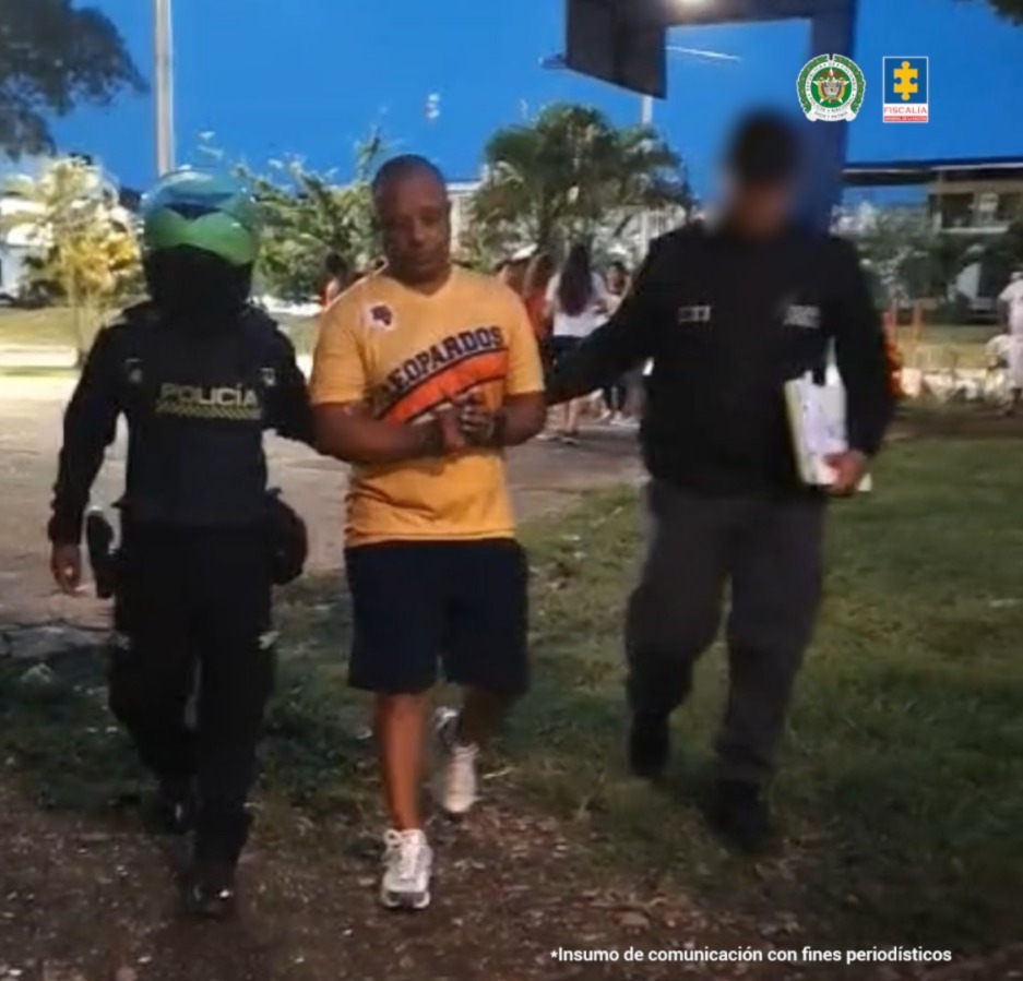 En la fotografía fue tomada en exteriores y se aprecia en acción de caminata, con los brazos adelante y esposado a Juan Carlos Reding Botero. Viste camiseta de color amarillo mostaza, de manga corta, con estampado diagonal de color negro y naranja; así como bermuda negra y zaptillas blancas. A su izquierda hay un servidor del CTI de la Fiscalía y a su derecha un uniformado de la Policía.