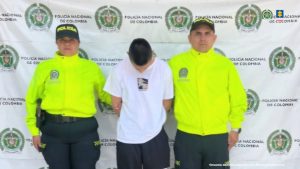 El capturado se encuentra en custodia por dos uniformados de la Policía Nacional. El capturado tiene un buzo blanco y detrás hay un backing de la Policía Nacional.