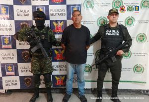El capturado se encuentra de frente y custodiado por un uniformado de la Policía y Ejército Nacional. Detrás hay un backing de la Policía y Ejército. El capturado tiene una camiseta de color negro jeans y tenis.